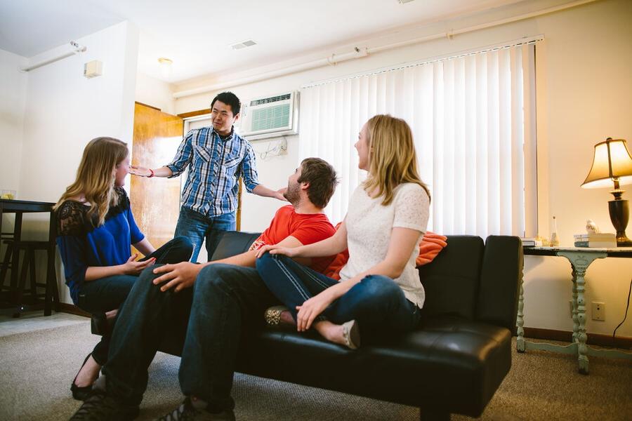 Man enters apartment into the living room with three people turned to talk to him.